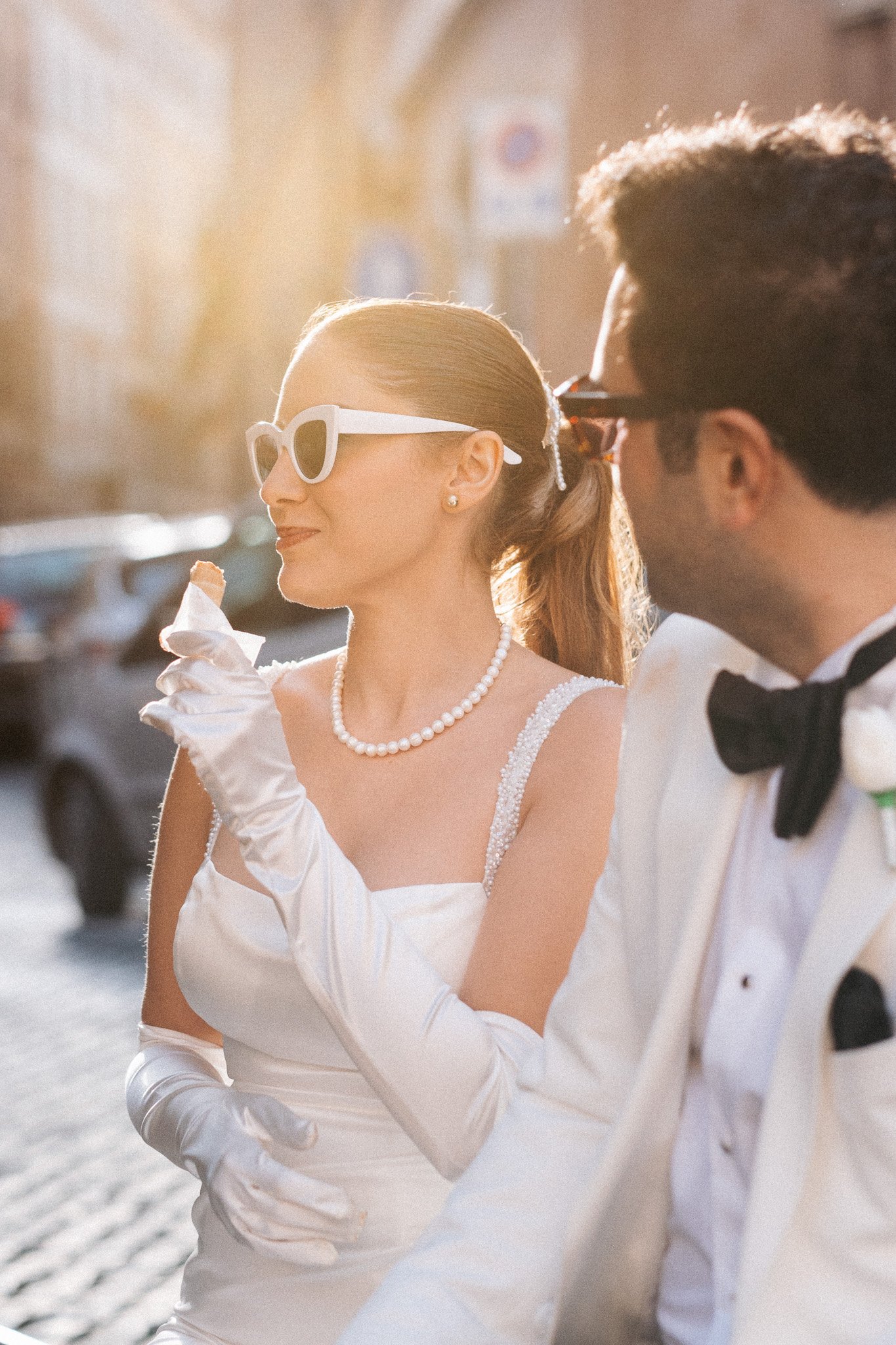 Bride in a white dress and gloves, with pearl necklace and white sunglasses, enjoying the last bite of gelato as sunlight glows behind her