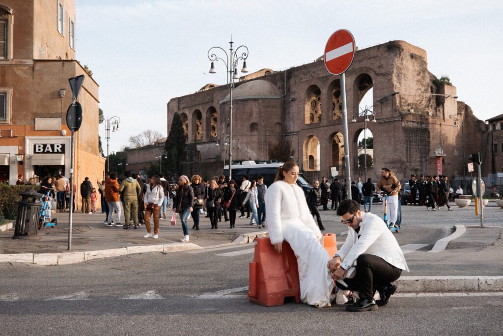 Groom kneeling to help his bride with her shoe as she sits on a street barrier, Roman ruins and city crowds in the background