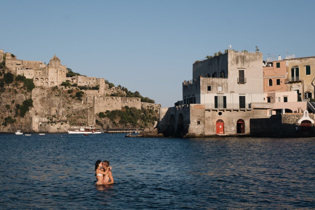 A couple embracing in the sea with Castello Aragonese and old waterfront buildings in the background in Ischia.