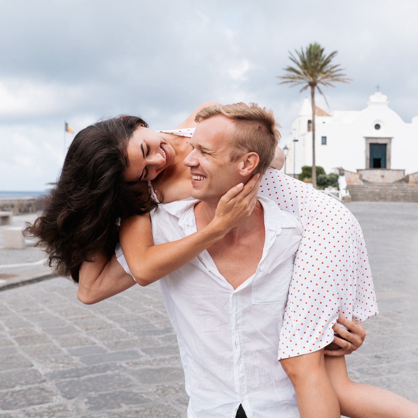 A smiling lady on a man’s back as they laugh together near a historic church with palm trees and stone pavement in Ischia.