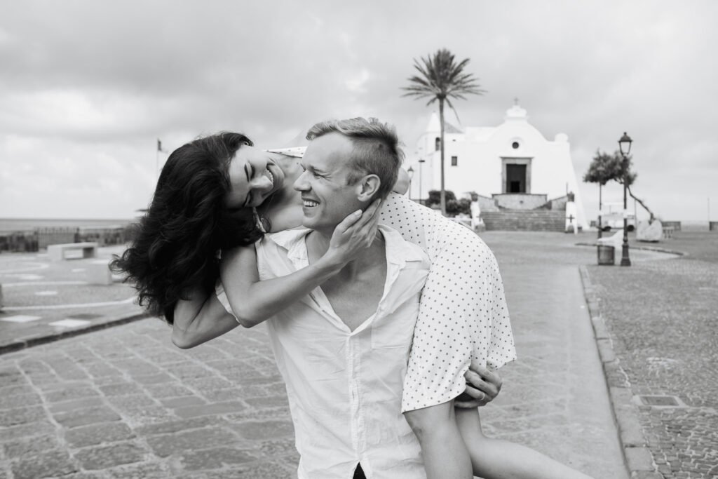 A smiling woman on a man’s back as they laugh together near a historic church with palm trees and stone pavement in Ischia, Italy.