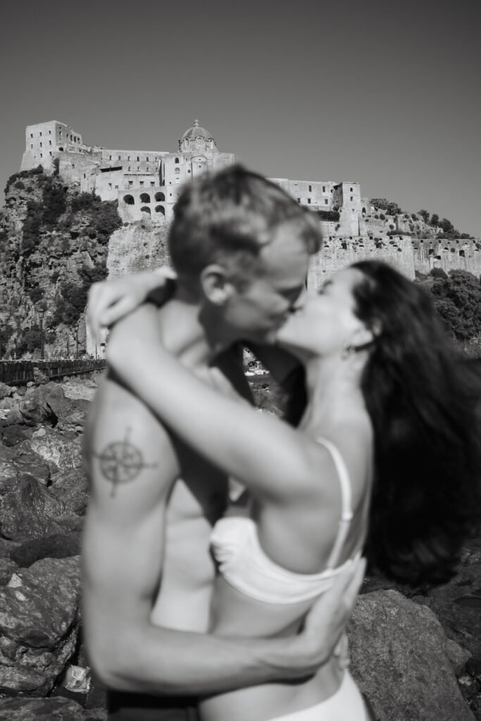 A couple kissing in swimwear, slightly blurred, with the Castello Aragonese clearly in focus behind them on a sunny day in Ischia.