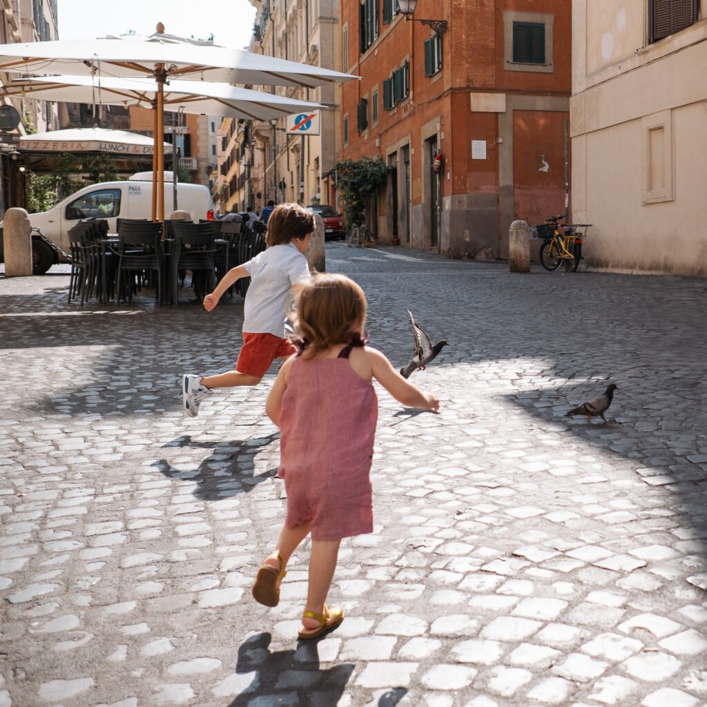 Two young children, a boy and a girl, run after pigeons on a cobblestone street in Rome. The scene is lit by afternoon sun, with colorful buildings, a pizzeria, and a bicycle in the background.