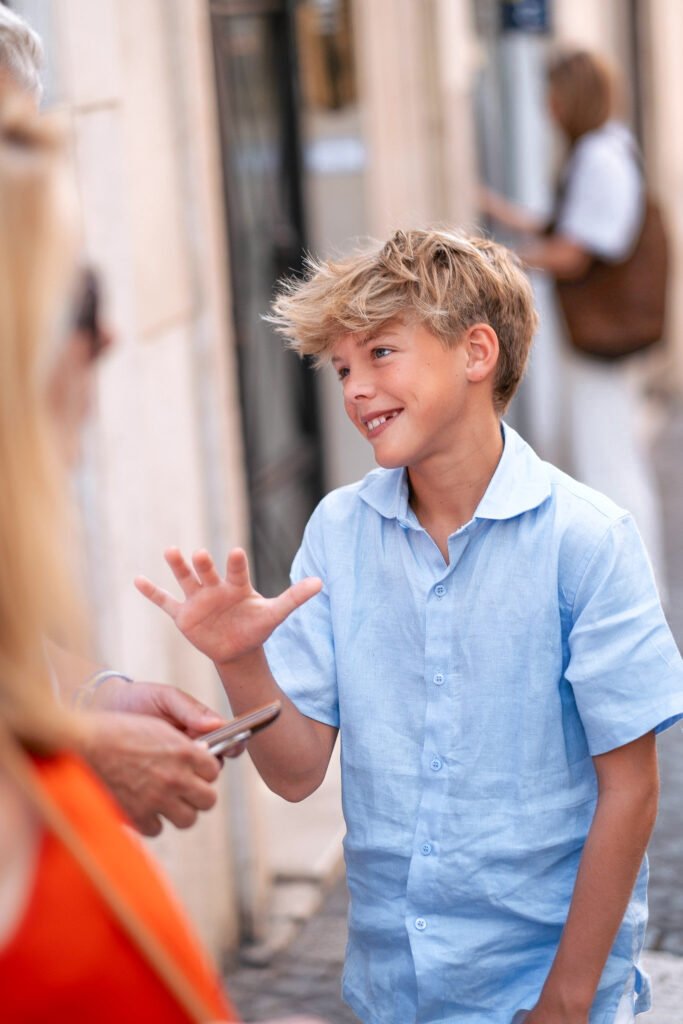 Smiling young boy in a light blue shirt holding up his hand mid-gesture while talking to adults during a casual family photoshoot on a cobbled street in Rome.