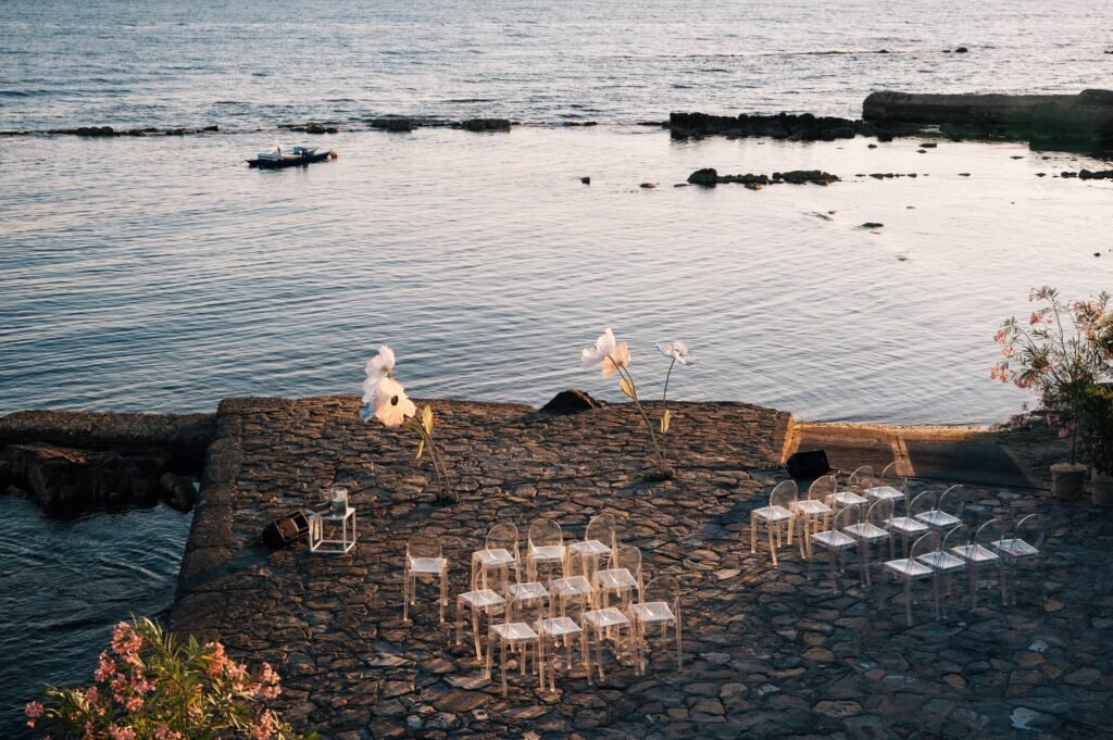 Seaside wedding ceremony setup at Villa La Saracena with ghost chairs and flowers.