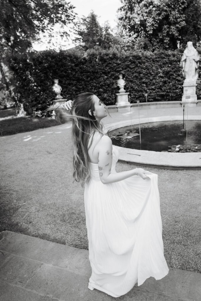 Woman in a white flowing dress standing beside a round fountain in an Italian garden, black-and-white engagement portrait in Rome, Italy.