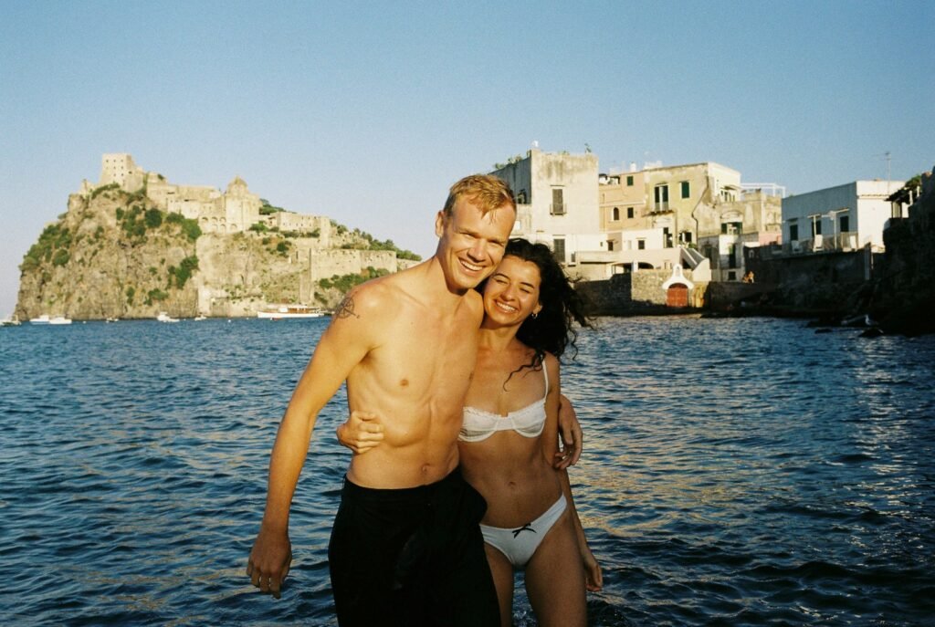 Smiling couple in swimwear embracing in the sea with an ancient Italian castle and coastal village in the background, perfect for romantic honeymoon photography in Rome and Italy.