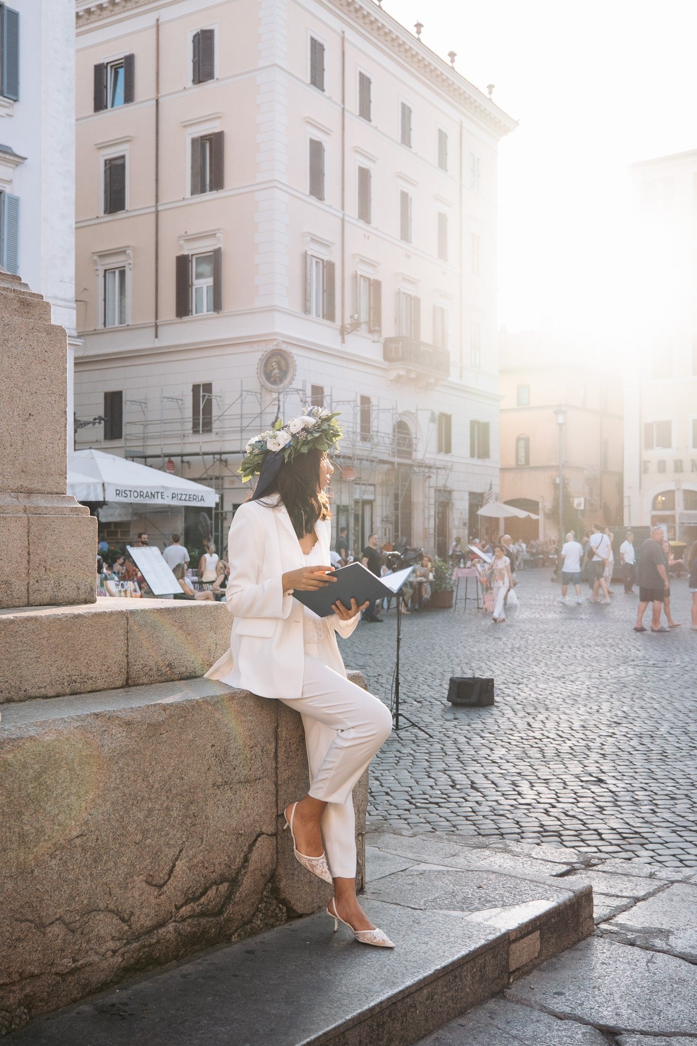 Woman seated on a stone monument, reading her diploma in a sunny square.