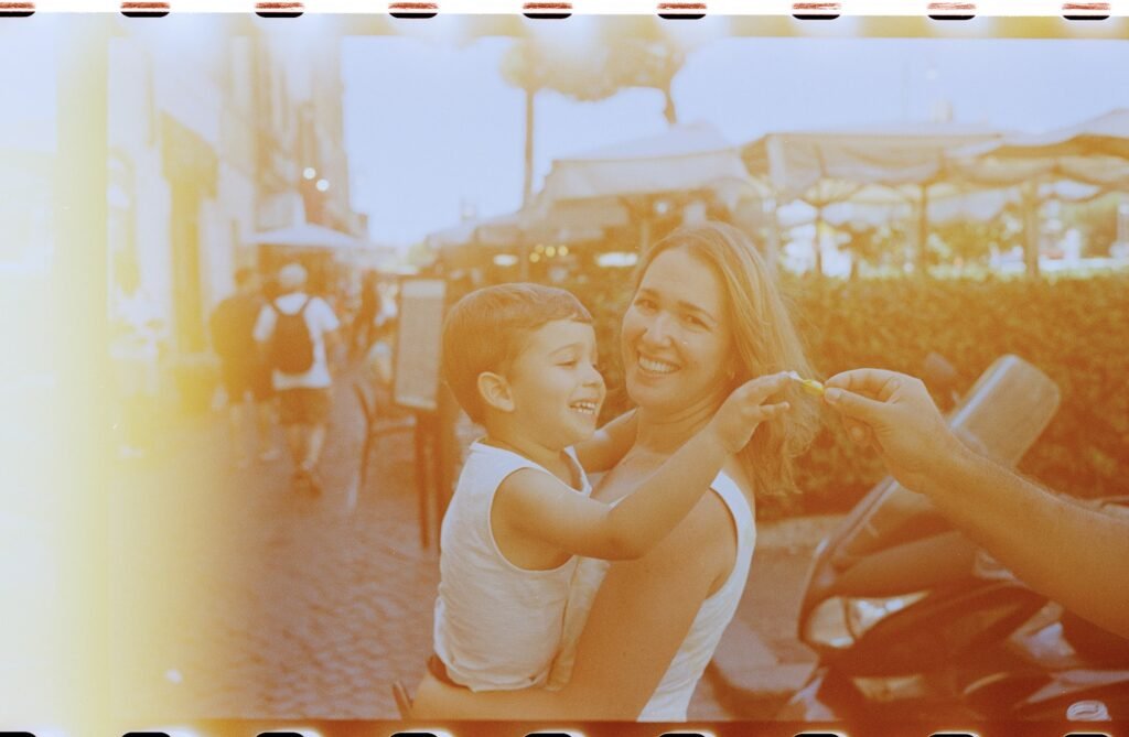 A smiling boy reaches for a candy while being held by his mother, with a restaurant scene behind them.