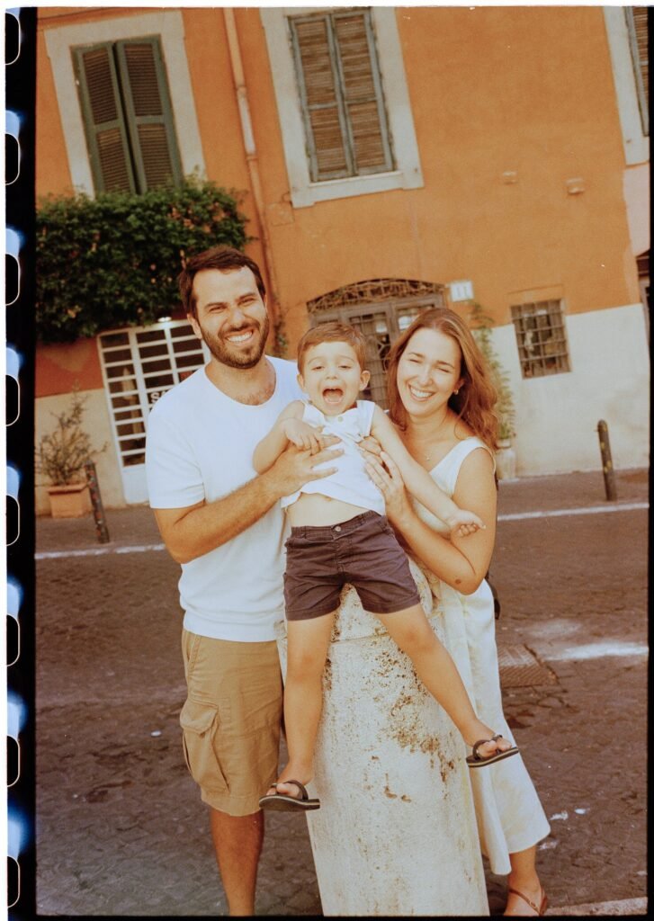 A smiling Brazilian family—father, mother, and young son—pose joyfully in front of an orange building in Rome.