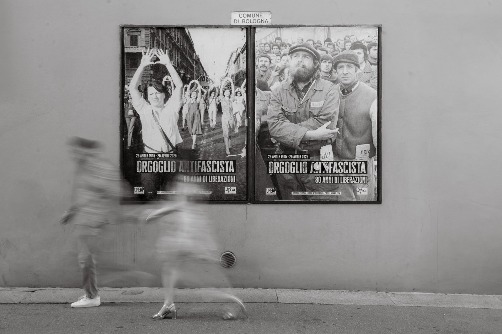 Blurred motion of a couple walking past antifascist posters in Bologna.