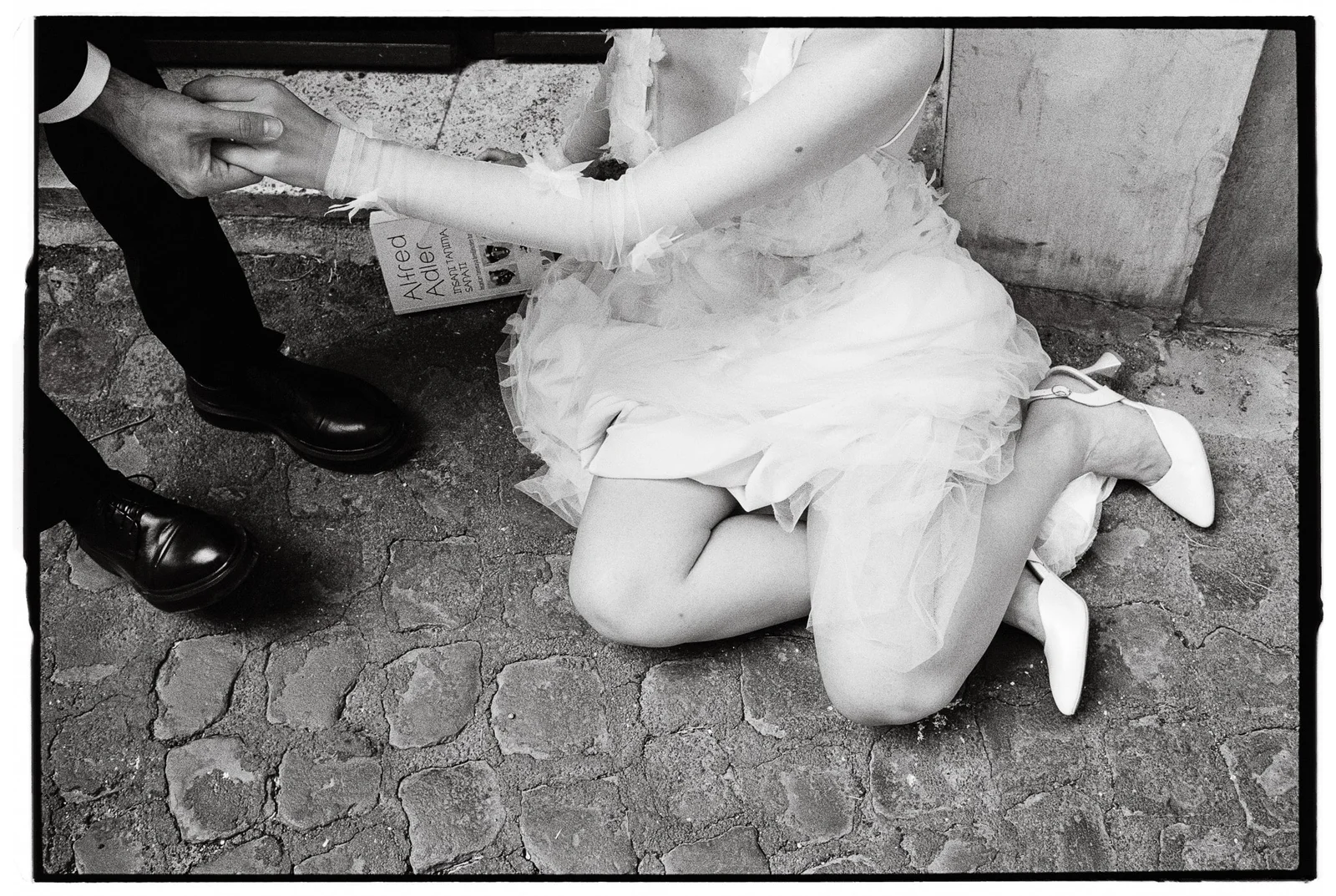 Bride kneeling in wedding gown holding groom’s hand on cobblestone street.