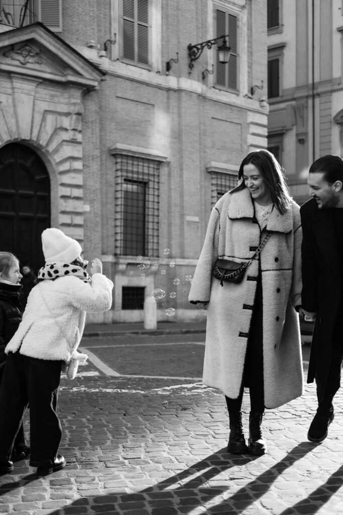 Coppia sorridente durante servizio fotografico a Roma mentre una bambina soffia bolle di sapone in una piazza storica.