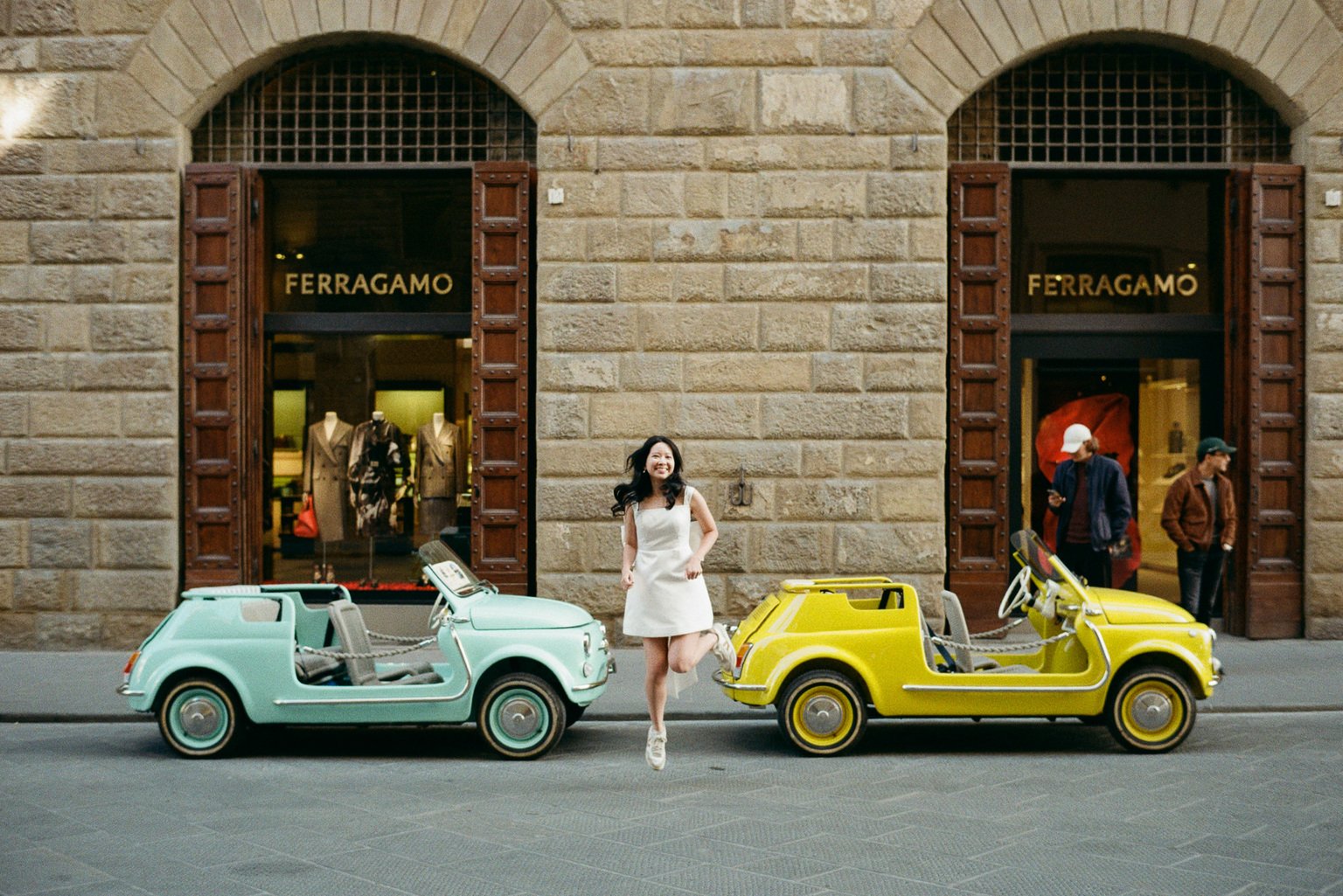 Film photograph of a woman in a white dress jumping between two vintage Fiat Jolly cars in front of the Ferragamo store in Florence.