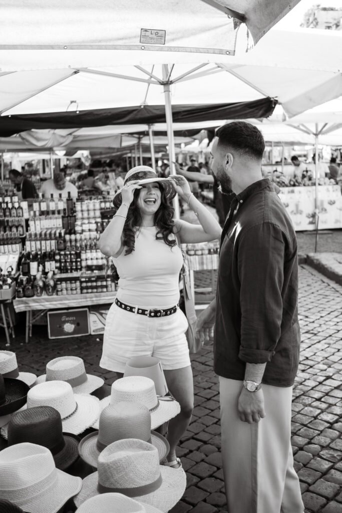 Candid black and white photo of a couple laughing and trying on hats at Campo de’ Fiori market during a couple photoshoot in Rome by photographer Gökhan Kutluer.