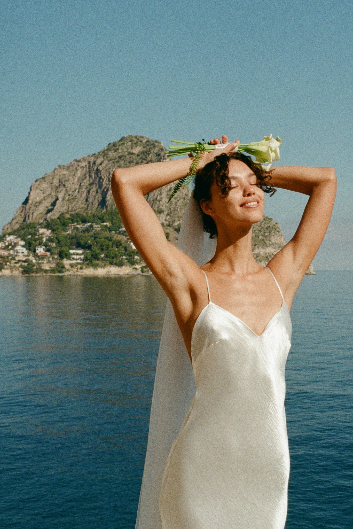 La sposa Désirée sorride al sole con un bouquet di calle davanti al mare di Mondello — fotografia di matrimonio su pellicola in Sicilia.