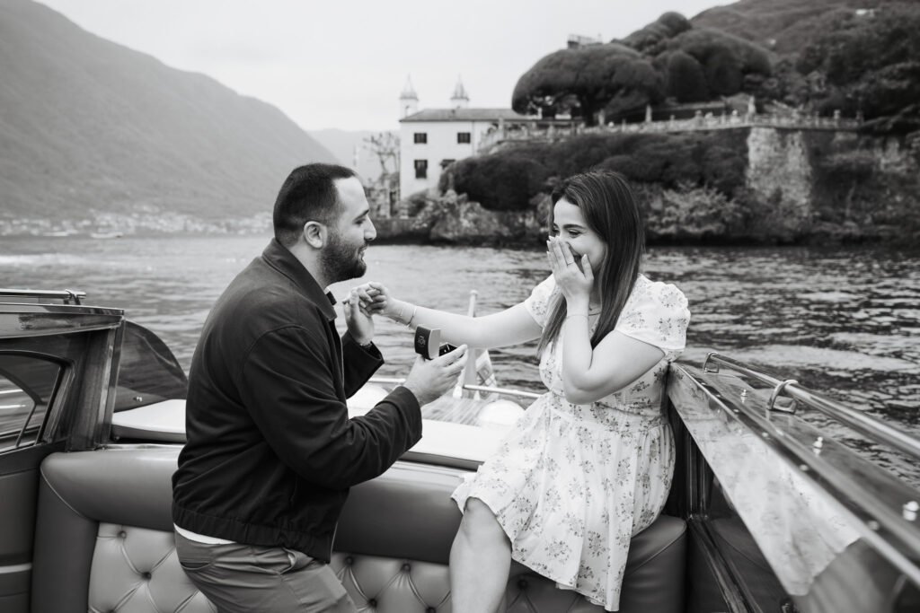 Black and white photo of a man on one knee proposing to a woman on a boat with a villa in the background.