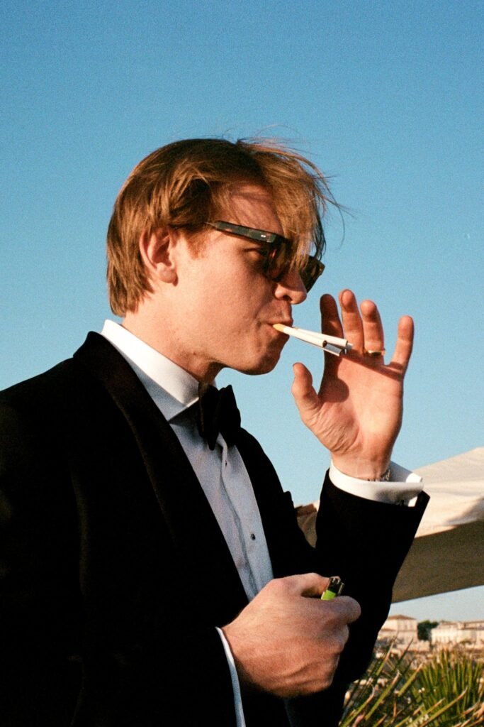 A close-up film photograph of a groom in a black tuxedo and sunglasses lighting two cigarettes simultaneously against a clear blue Italian sky. Shot by a documentary wedding photographer using a 35mm Contax G2 camera.