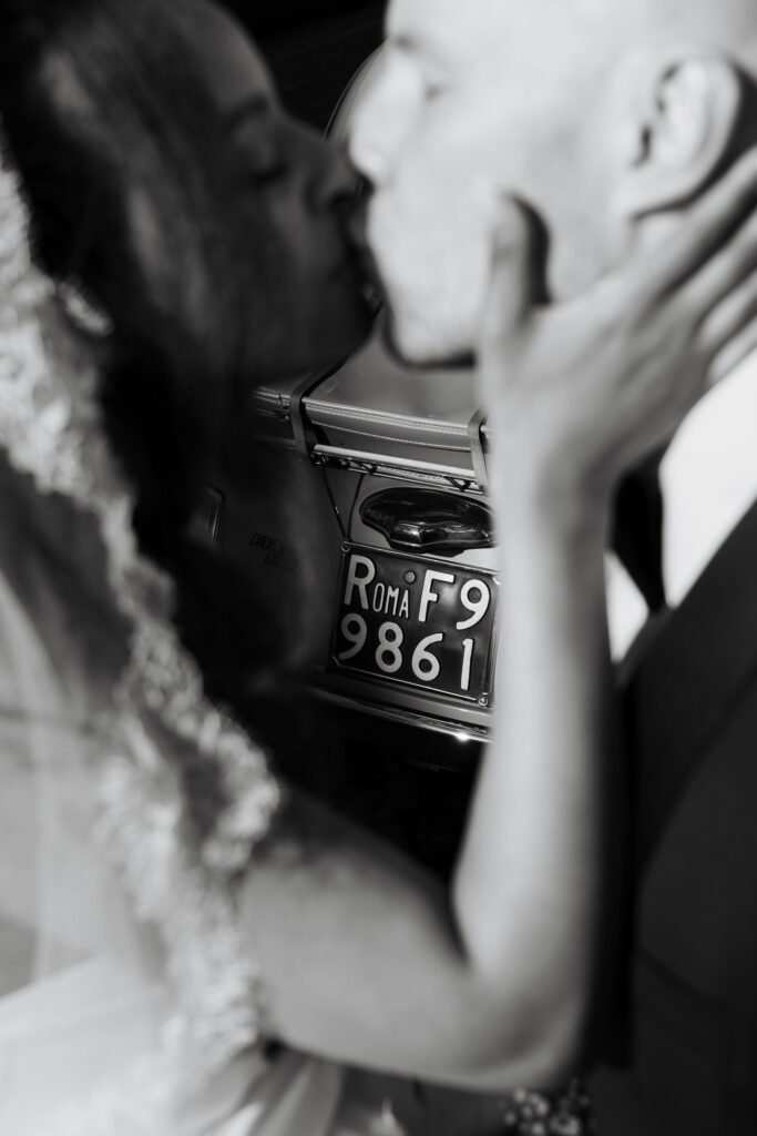 Black and white close-up of bride and groom kissing beside a vintage Fiat 500 after their micro wedding in Rome.