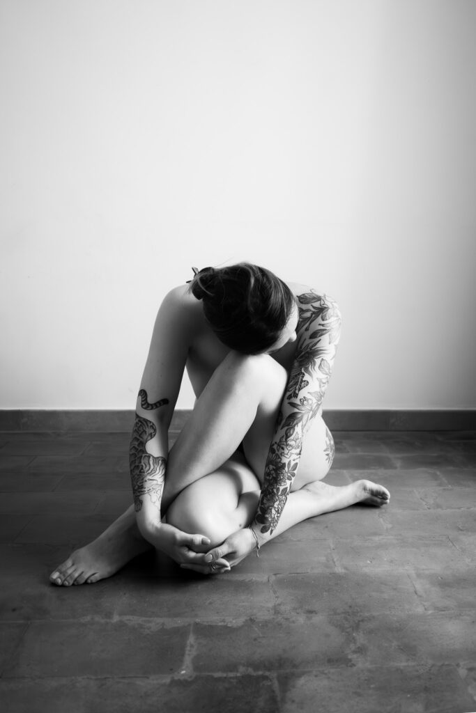 Intimate black and white fine art nude portrait of a woman curled on a stone floor in Rome, hugging her knees, with extensive tattoos visible on her arms and side. Artistic nude photography Italy.