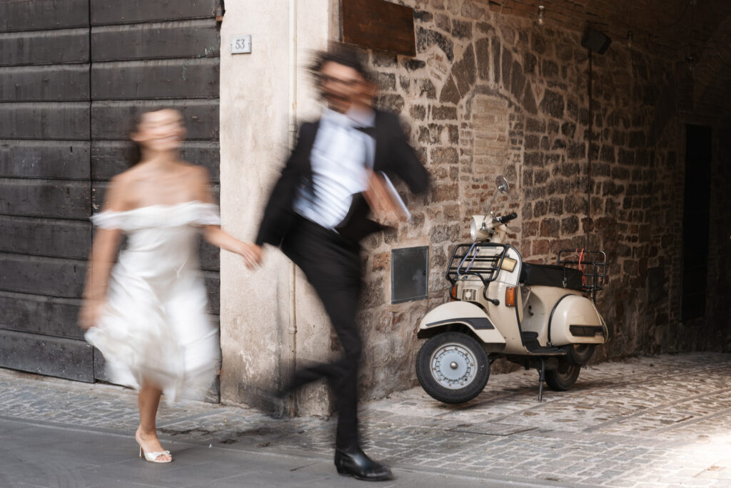 Bride and groom running hand in hand past a vintage Vespa in Spello during a playful elopement photography session in Umbria.
