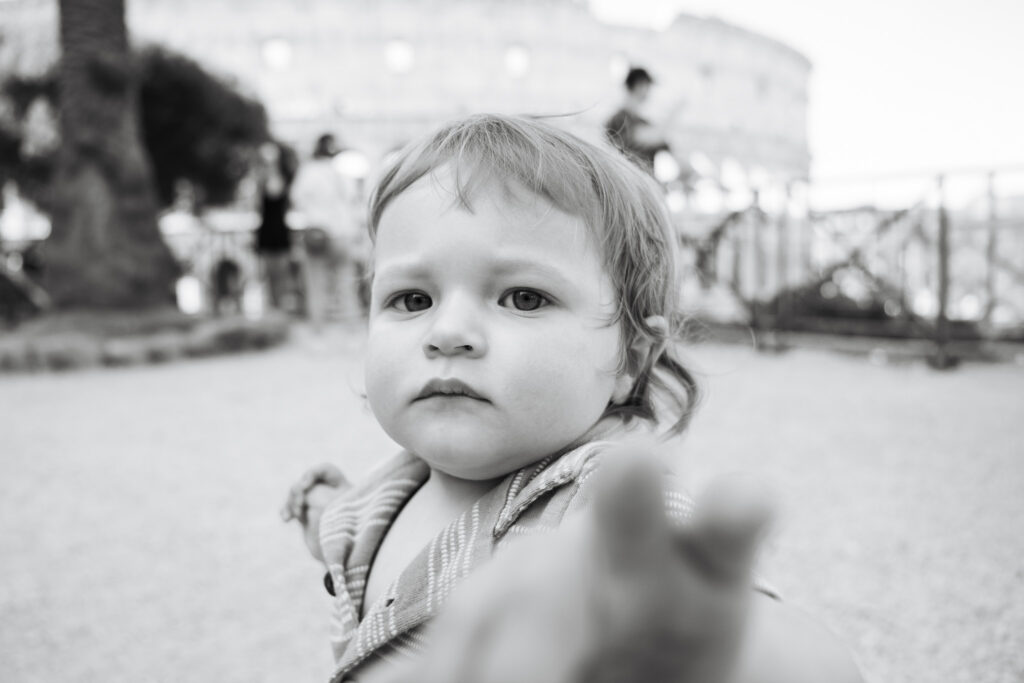 Toddler reaching toward the camera during a candid family photoshoot in Rome with the Colosseum softly blurred in the background.