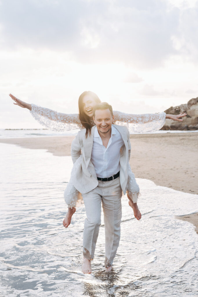 Bride riding on the groom’s back during a playful sunset wedding photoshoot on Anzio beach near Rome.