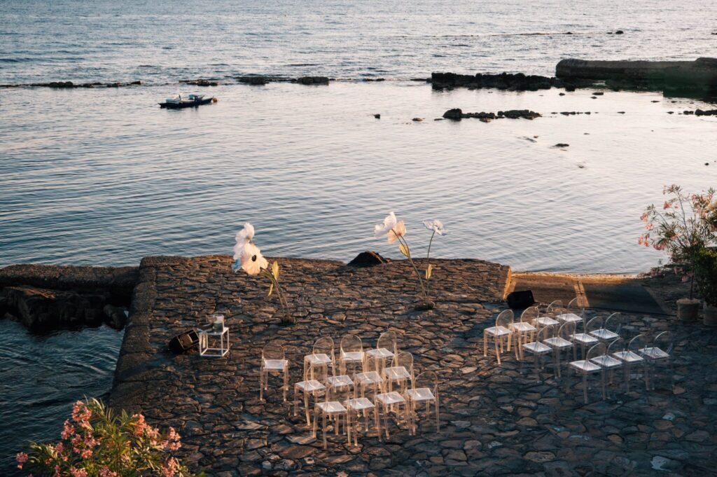 Sedie trasparenti e composizioni floreali preparate su una terrazza in pietra affacciata sul mare.