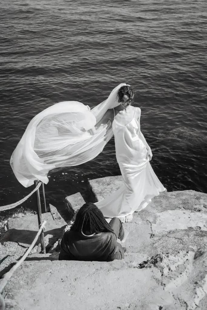Smiling bride holding her long veil as it blows in the wind beside the Mediterranean sea.