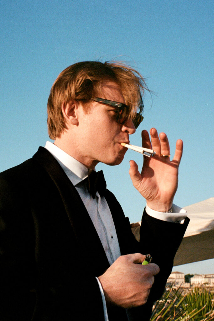 Invitato in smoking e occhiali da sole mentre accende una sigaretta, fotografato su pellicola 35mm.