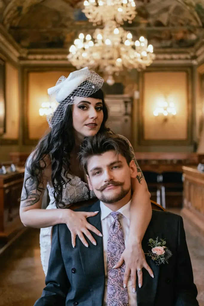 Sposa abbracciata allo sposo seduto sotto grandi lampadari durante il loro matrimonio a Bologna.