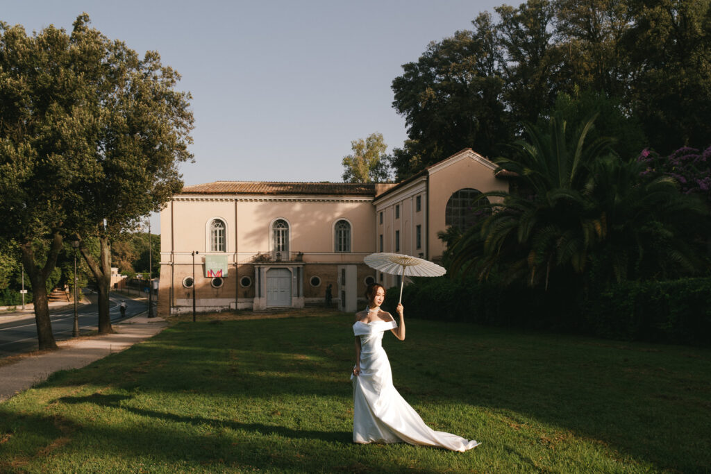 Japanese bride from Tokyo in a white wedding dress holding a parasol in the evening light at Villa Borghese during a pre wedding photoshoot in Rome, Italy.