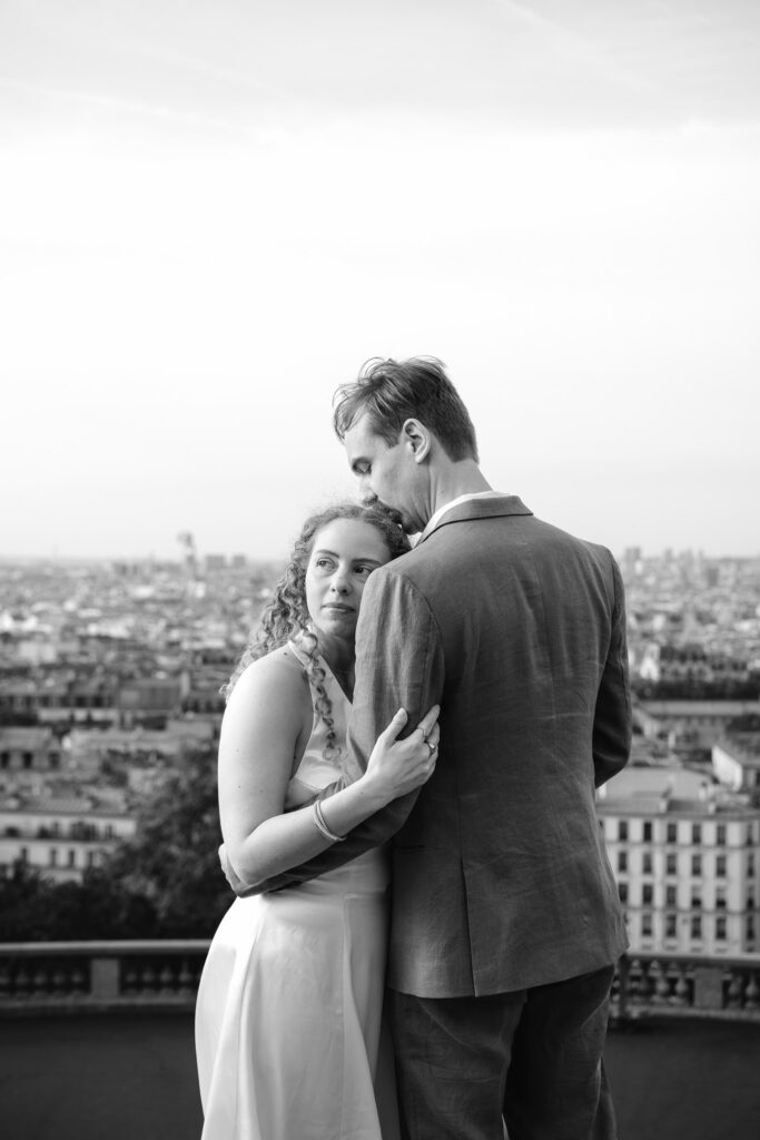 Black and white portrait of Kaylyn and Rynhardt embracing with a panoramic view over Paris during their Montmartre couple photoshoot.
