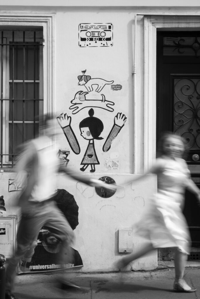 Black and white motion blur photograph of Kaylyn and Rynhardt walking past street art during their Montmartre couple photoshoot in Paris.
