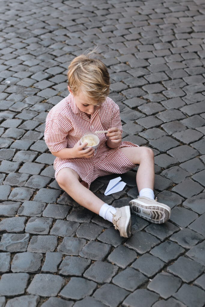 Child sitting on a cobblestone street eating gelato during a family photoshoot in Rome