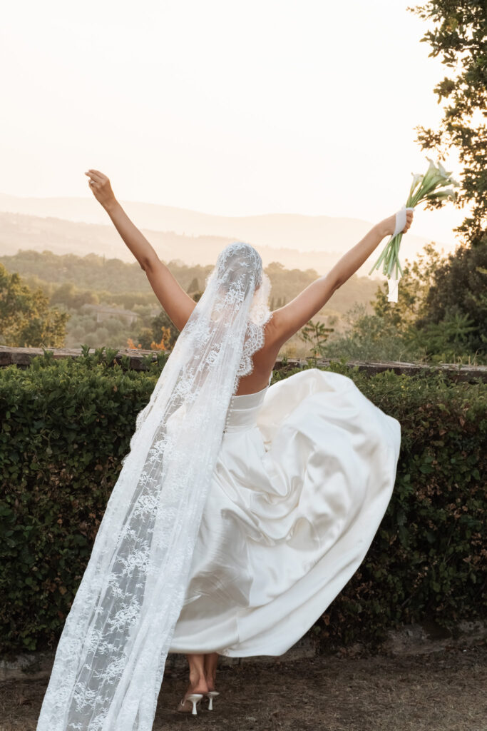 Bride seen from behind with arms raised, holding her bouquet and looking over the Tuscan countryside at golden hour.