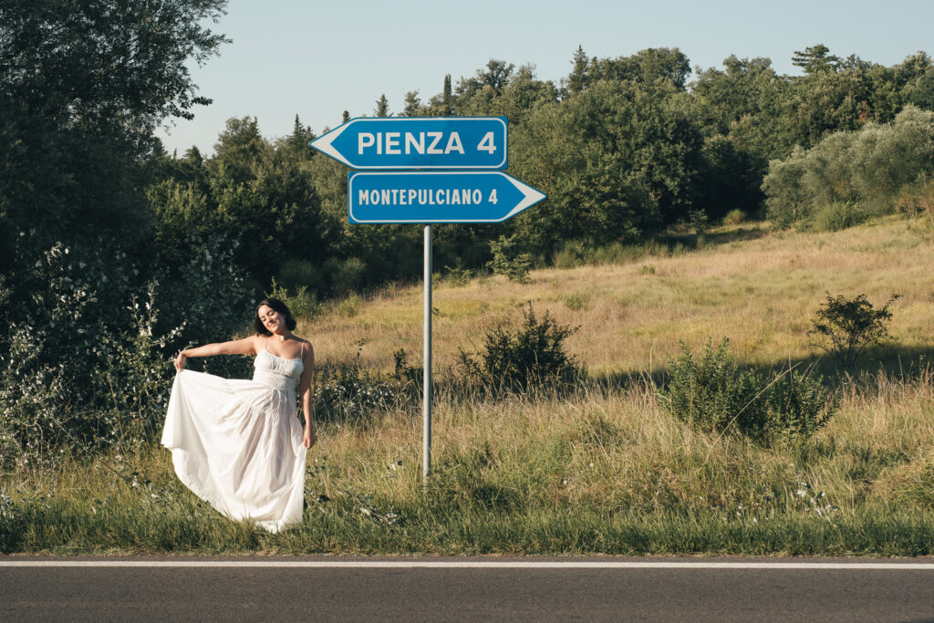 Bride posing beside a road sign pointing to Pienza and Montepulciano during a Tuscany elopement in Val d’Orcia.