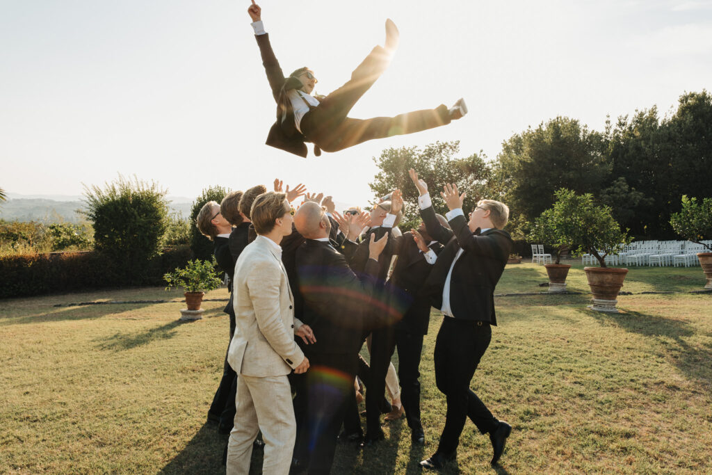 Sposo lanciato in aria dagli amici durante una festa di matrimonio all’aperto nella campagna toscana.