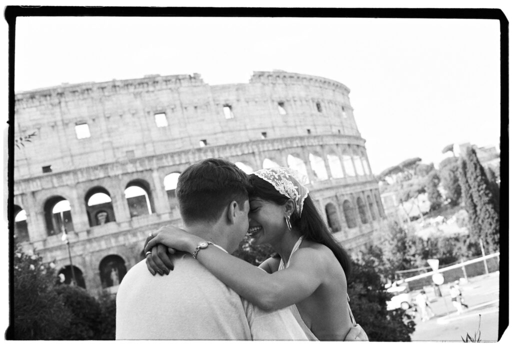 Aditi e il suo compagno si abbracciano davanti al Colosseo durante un servizio fotografico di fidanzamento a Roma su pellicola in bianco e nero.