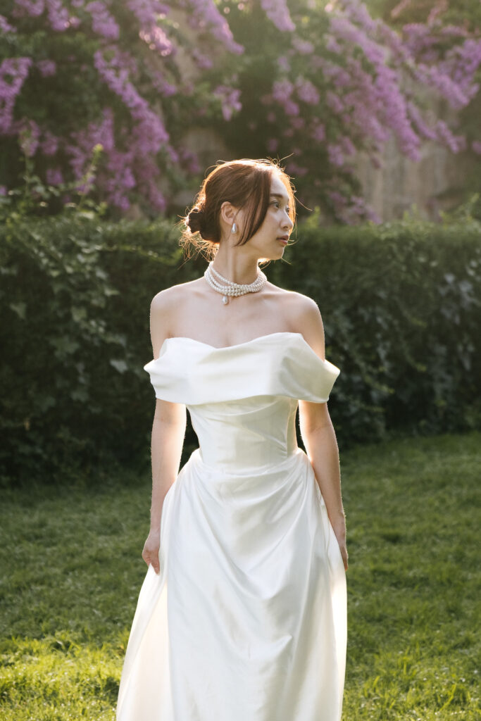 Ritratto di Kanaka in abito bianco nella luce dorata di Villa Borghese, circondata dal verde e da fiori viola durante un servizio prematrimoniale a Roma.