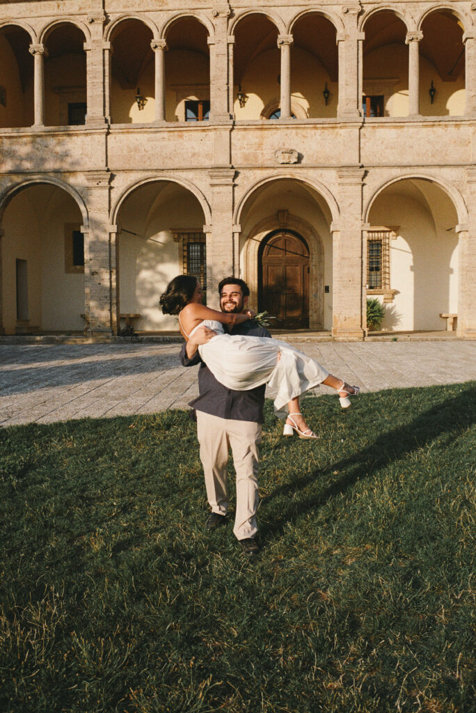 Groom carrying the bride in front of historic Tuscan architecture during their 35mm film elopement in Val d’Orcia.