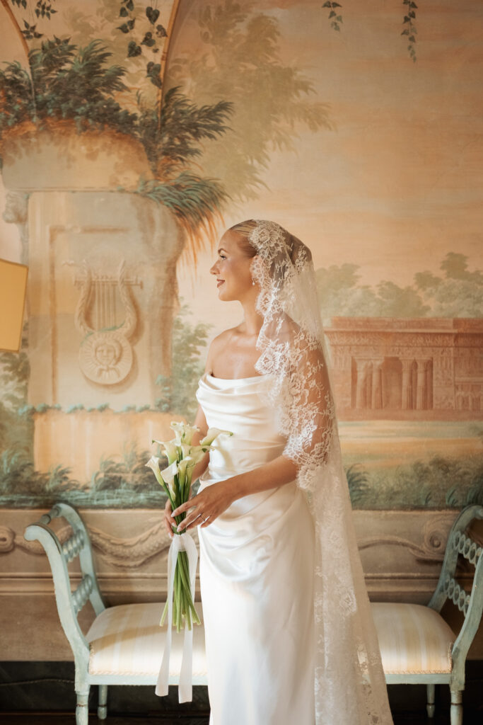 Bride wearing a lace veil and holding calla lilies inside the historic interiors of Villa Catignano in Tuscany.