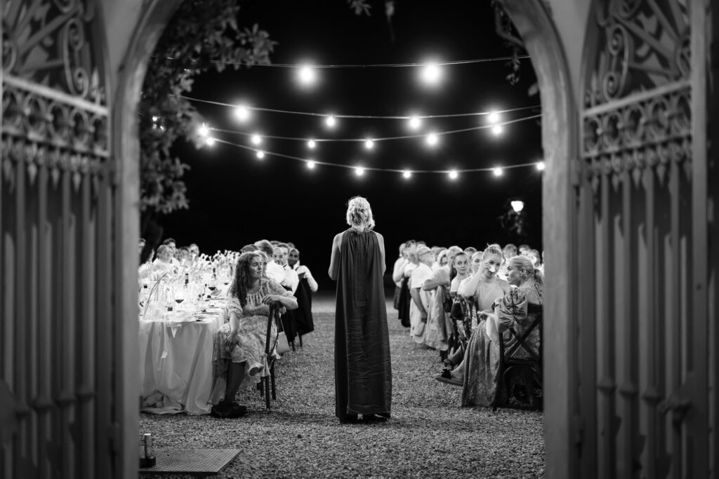 Black-and-white photograph of an evening wedding speech between long dinner tables under string lights at Villa Catignano.
