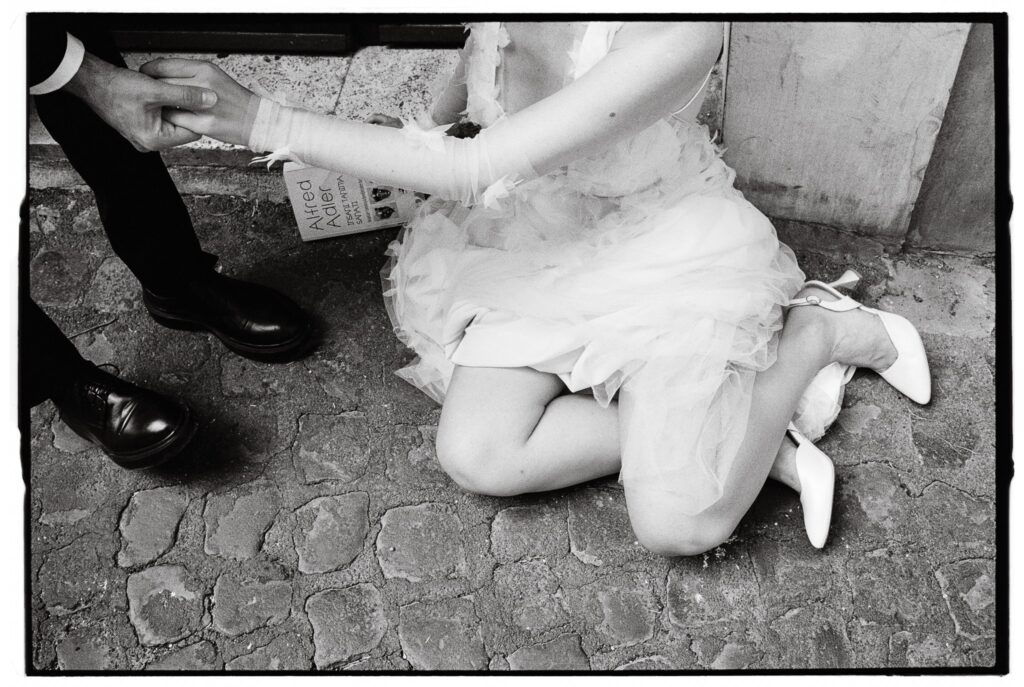 Bride in a tulle wedding dress sitting on cobblestones while holding her partner's hands, captured on black-and-white 35mm film.