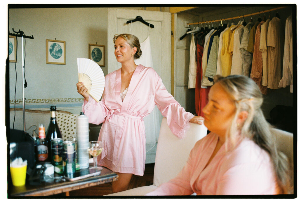 Bride in a pink satin robe holding a white folding fan while getting ready with another woman, photographed on 35mm film.