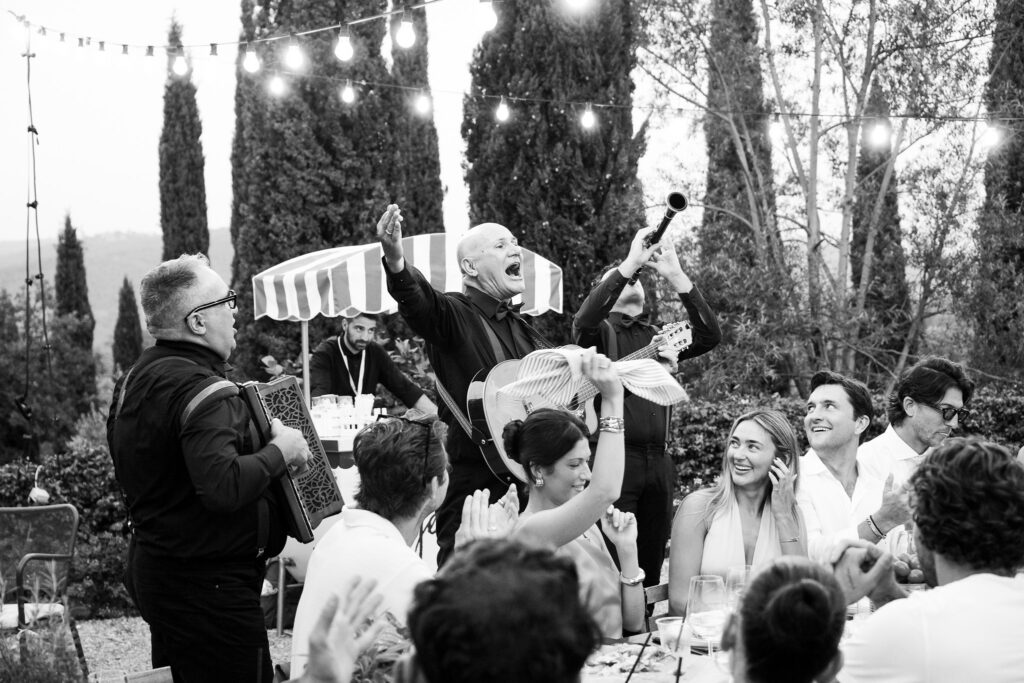 Wedding guests laughing and celebrating around outdoor dinner tables as live musicians play accordion and guitar beneath string lights.