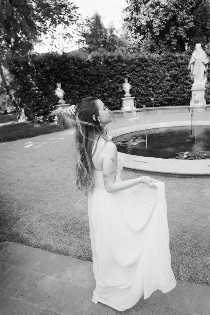 Bride in a flowing white dress standing beside a garden fountain, touching her hair in a black-and-white wedding portrait.