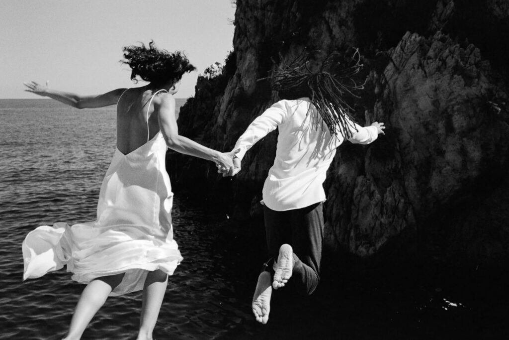 Newlyweds jumping hand in hand from a rocky ledge into the sea, the bride's white wedding dress flowing through the air in a black-and-white photograph.