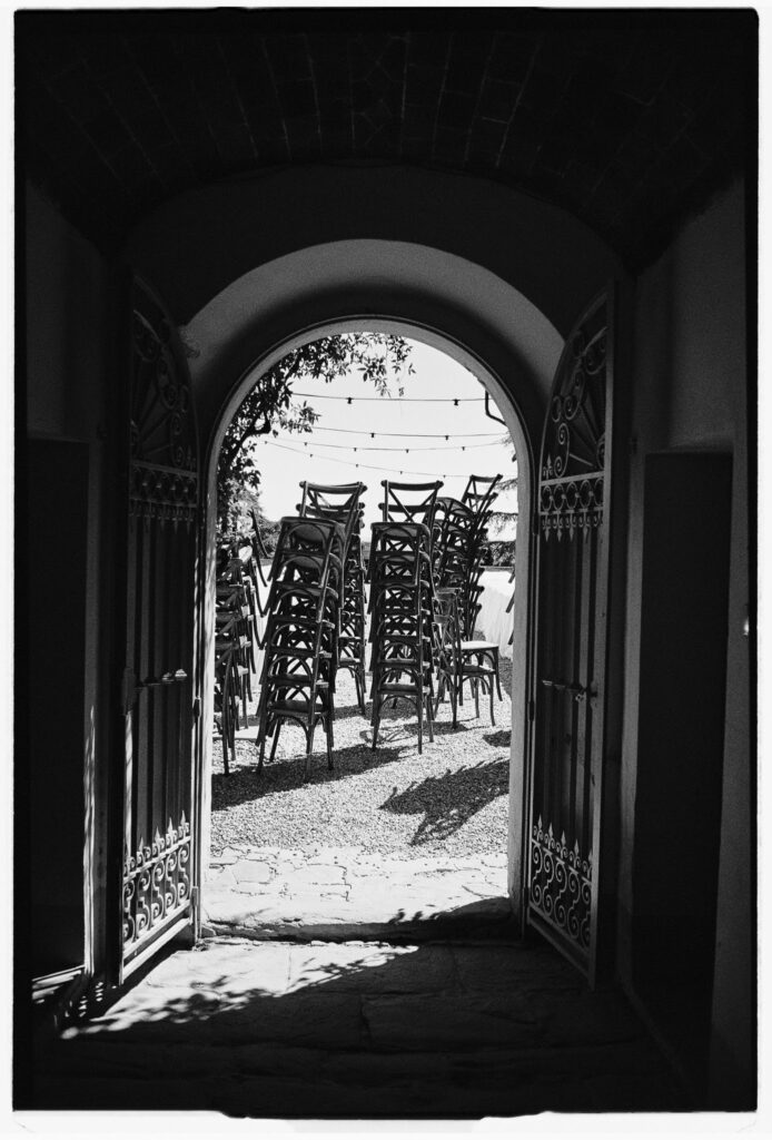 Black-and-white 35mm photograph of stacked wedding chairs seen through an arched doorway at a wedding venue.