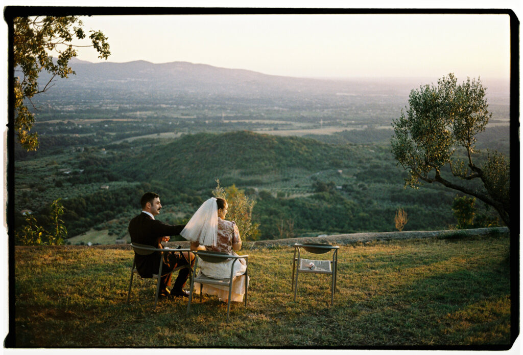 Newlyweds sitting together on chairs overlooking a green valley at sunset, photographed on 35mm analogue film.